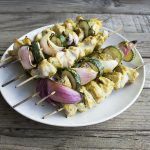 a stack of kabobs with chicken, zucchinis, red onions on a white plate on a wooden board