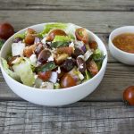 A white ceramic bowl with the salad inside and a small white ceramic bowl next to it with dressing on a wood board with tomatoes