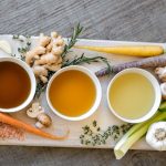 Three bowls of different broths with veggies around them on a cutting board