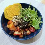 A dark blue bowl filled with roasted yellow and red beets, carrots, fresh greens, and quinoa