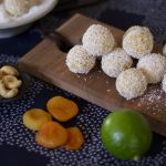 Sunshine bites rolled with a sprinkle of coconut on top. They are stacked on a cutting board with lime, dried apricots and cashews next to the balls