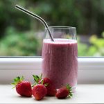 a glass with a pink smoothie with a metal straw. Strawberries around it in front of a window