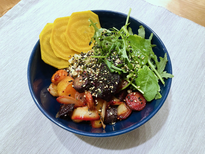A dark blue bowl filled with roasted yellow and red beets, carrots, fresh greens, and quinoa