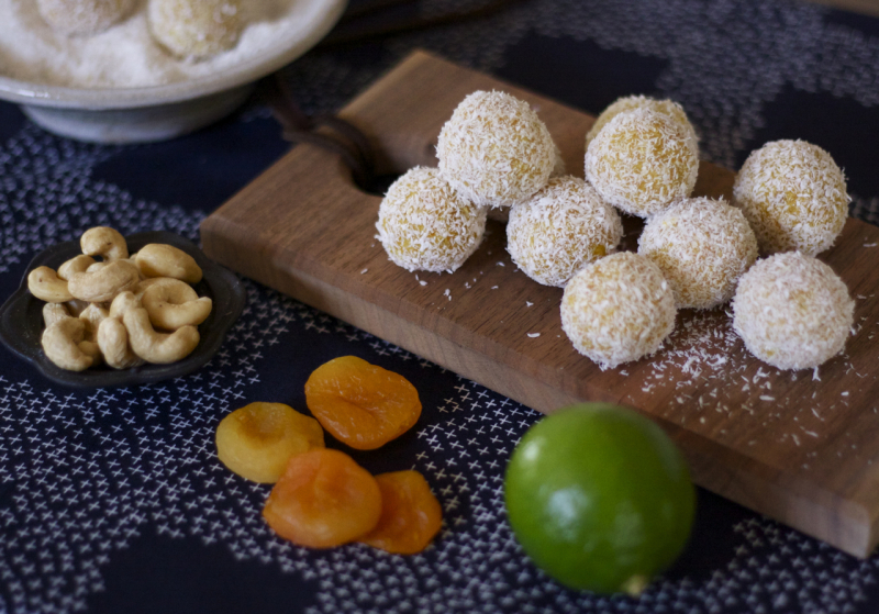 Sunshine bites rolled with a sprinkle of coconut on top. They are stacked on a cutting board with lime, dried apricots and cashews next to the balls