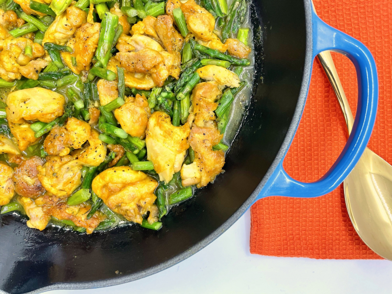 A blue skillet filled with turmeric-seasoned chicken and green asparagus with a bright red napkin