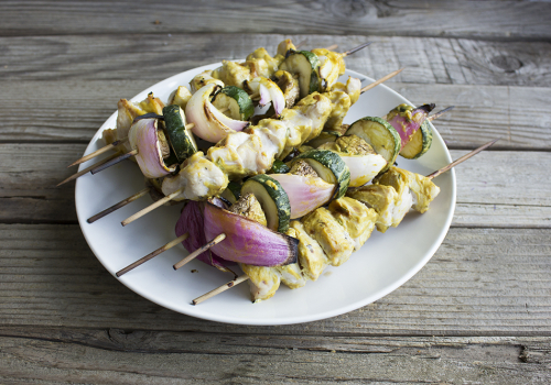 a stack of kabobs with chicken, zucchinis, red onions on a white plate on a wooden board