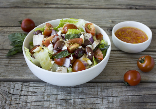 A white ceramic bowl with the salad inside and a small white ceramic bowl next to it with dressing on a wood board with tomatoes