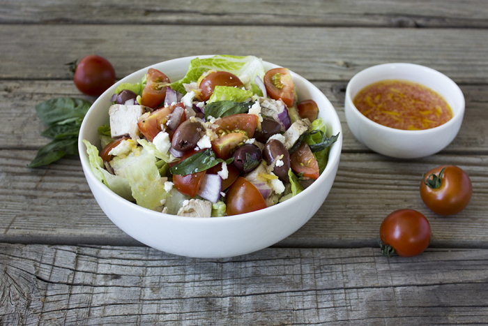 A white ceramic bowl with the salad inside and a small white ceramic bowl next to it with dressing on a wood board with tomatoes